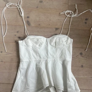 Vit korsett-topp från Zara - Supersnygg vit korsett-topp från Zara med tunna knytband och formade kupor. Toppen har en lätt utsvängd nederdel och smockad rygg för extra komfort. 