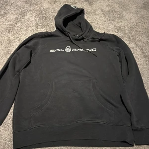 Svart Sail Racing hoodie med tryck - Svart hoodie från Sail Racing med vit logotyp på bröstet och en mindre logga på huvan. Tröjan har klassisk känguruficka, snörning i huvan och långa ärmar. Perfekt för chill dagar och streetwear-stil. Materialet är mjuk bomull som känns skönt mot huden.