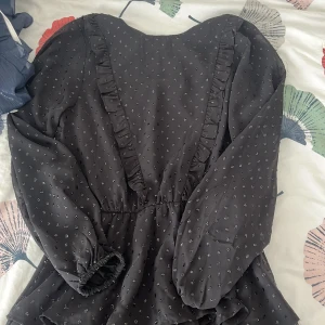 Svart kort klänning med volang från Gina Tricot - Supersöt svart kort klänning från Gina Tricot med små glittriga prickar och volangdetaljer framtill. Klänningen har långärmade puffärmar med resår vid handlederna och ett lager på kjolen som ger extra volym. Perfekt för dig som gillar en feminin och trendig look. Priset kan även diskuteras