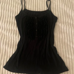 Svart linnetopp med volang och knappar - Svart linnetopp med smala axelband och snygga volangdetaljer framtill. Fyra svarta knappar pryder mitten av toppen och ger en cool touch. Materialet känns mjukt och lätt, perfekt för varma dagar. Passformen är figurnära och toppen är ärmlös.
