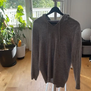 Grå hoodie med huva och snörning - Mysig grå hoodie med huva och snörning i halsen. Tröjan har långärmade ärmar och är tillverkad i ett mjukt stickat material som känns skönt mot huden. Perfekt för chill dagar och enkel att matcha med jeans.