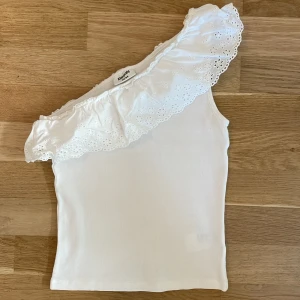 Vit oneshoulder topp med volang Kappahl 158/164 - Supersöt vit topp från Kappahl i oneshoulder-modell med bred broderad volang längs halsen. Toppen är ärmlös och har en ribbad struktur i tyget. 