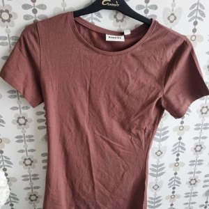 Brun t-shirt från Kappahl - En enkel brun t-shirt från Kappahl med rund hals och korta ärmar. Modellen är klassisk och passar dig som gillar en clean och chill stil. Materialet är mjuk bomull som känns skönt mot huden. Perfekt att matcha med jeans eller shorts.