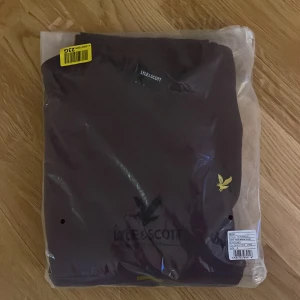 Vinröd tröja från Lyle & Scott - Snygg vinröd långärmad tröja från Lyle & Scott med klassisk rund hals och gul broderad logga på bröstet. Tröjan är i mjuk bomull och har en stilren, normal passform som funkar till jeans eller chinos. Perfekt för dig som gillar enkel och clean stil.