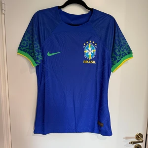 Blå Brasil fotbollströja - Säljer en officiell Brasilien fotbollströja från Nike i blått med grönt Nike-logga och gult Brasil-tryck. Tröjan har korta ärmar med leopardmönster i grönt och blått samt gula detaljer vid ärmslut. Materialet är lätt och andas, Dri-Fit ADV för extra komfort under match.
