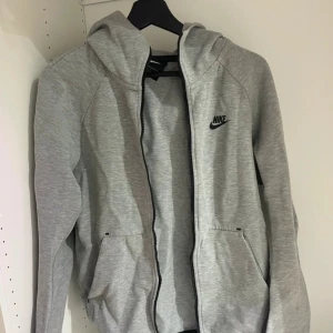 Grå hoodiejacka från Nike - Snygg grå hoodiejacka från Nike med dragkedja och huva. Jackan har två praktiska fickor framtill och en stilren Nike-logga på bröstet. Perfekt för chill dagar eller när du vill ha en sportig look. Tillverkad i mjuk bomullsmix för skön komfort.