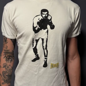 Vit t-shirt med boxartryck från Ingo Boxers - Säljer en vit t-shirt från Ingo Boxers i storlek L. Framsidan har ett stort svart tryck med en boxare och en gul Ingo Boxers-logga. T-shirten har rund hals och korta ärmar, tillverkad i tjock och mjuk bomull som känns bekväm mot huden. Modellen på bilden är 190 cm lång och bär storlek L – men passformen motsvarar mer en M. Perfekt för dig som gillar sportiga och retro vibbar.