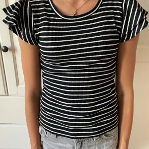 Randig svartvit topp från Lindex - Snygg ribbad topp från Lindex med svart och vita horisontella ränder. Modellen har korta volangärmar och rund halsringning. Materialet är mjukt och stretchigt, perfekt för dig som gillar bekväma och stilrena plagg. Passar dig som vill ha en enkel men cool look.