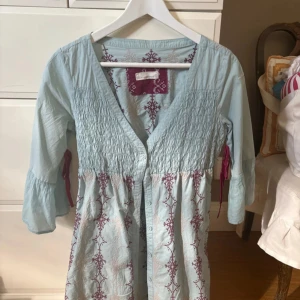 Odd Molly ljusblå broderad klänning - Supersöt ljusblå kortklänning från Odd Molly med broderade mönster i vinrött och beige. Klänningen har v-ringning, knappar framtill, trekvartsärm med volang och dekorativa knytband i vinrött vid ärmarna. Perfekt för dig som gillar boho-vibe och detaljer.