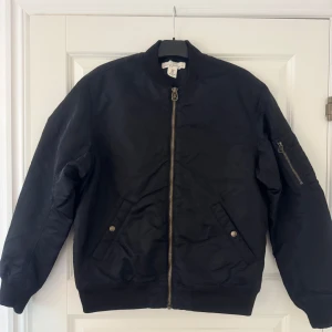 Svart bomberjacka i medium - Svart bomberjacka från Label of Graded Goods i storlek medium. Jackan har klassisk ribbad krage, dragkedja framtill och fickor med tryckknappar. Ärmarna är långärmade och har en extra ficka med dragkedja på ena armen. Perfekt för en cool streetstil.