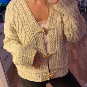 Beige stickad kofta med träknappar - Supermysig beige stickad kofta med grova kabelstickade detaljer och snygga träknappar framtill. Koftan har långärmade ärmar och en loose passform som ger en chill vibe. Perfekt att slänga på sig när det är kallt ute och du vill vara både varm och snygg.