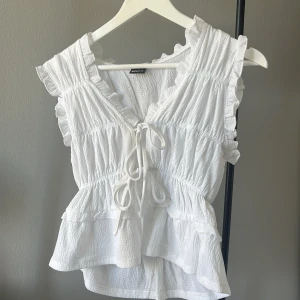 Vit blus med volanger och knyt - Supersöt vit blus från Gina Tricot med volangdetaljer och v-ringning. Blusen har knyt framtill, är ärmlös och har en lätt rynkad struktur i tyget. Passar till vad som helt 💞