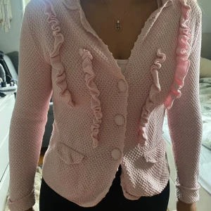 Rosa stickad kofta Odd Molly superkvalitet Medium/small - Säljer en ljusrosa cardigan/kofta från Odd Molly med snygga volangdetaljer framtill och klädda knappar. Den är stickad i 100% bomull och har broderad logga på ryggen. Perfekt för dig som gillar en söt och unik stil. Väldigt fin kvalitet. Något mindre än medium märkt 2. Den har också ett skärp som man kan knyta runt midjan eller som detalj på baksidan. Ena hylsan är dock trasig, går att laga. Se bild 5.