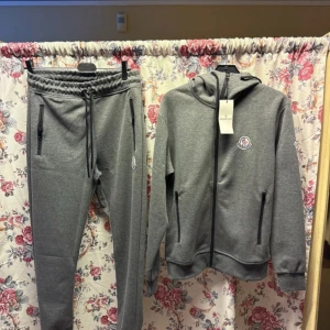 Grå Moncler hoodie med dragkedja - Säljer en grå hoodie från Moncler med huva och hel dragkedja framtill. Tröjan har Moncler-logga på bröstet, svarta dragkedjor på fickorna och ribbade muddar. Materialet är mjuk bomull och polyester, perfekt för chill dagar. Snygg och stilren design som funkar till allt.