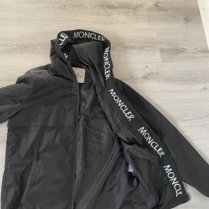 Svart vindjacka från Moncler - Svart vindjacka från Moncler med huva och vit logotyptext längs kanten. Jackan har dragkedja framtill, två sidofickor med dragkedja och en stilren Moncler-logga på bröstet. Materialet är lätt och syntetiskt, perfekt för blåsiga dagar.