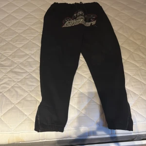 Svarta mjukisbyxor Trapstar London Shooters - Svarta mjukisbyxor från Trapstar med broderad London Shooters-logga och pantermotiv framtill. Byxorna har snörning i midjan, elastiska benslut och dragkedjor vid anklarna. Perfekt för dig som gillar streetwear och vill sticka ut med snygga detaljer.