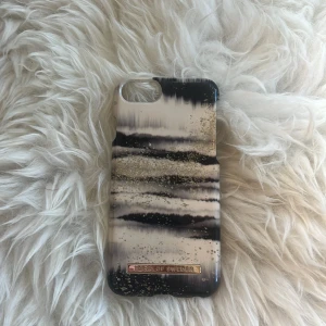 Randig mobilväska från iDeal of Sweden - För iPhone 6s,7 och 8. Snyggt mobilskal med randigt mönster i svart, beige och vitt, dekorerad med guldglittriga detaljer. Stilren design med ett metallmärke längst ner. Passar perfekt för dig som vill ha en unik och trendig look på mobilen.💓