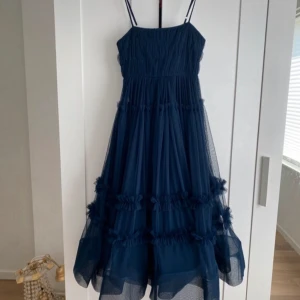 Marinblå långklänning med volanger - Säljer en marinblå långklänning med tunna axelband och flera lager av mjuk tyll. Klänningen har volangdetaljer längs kjolen som ger extra fluff och rörelse. Helt ny ordinarie pris= 1000 kr! Har tyvärr ingen prislapp men har aldrig används.