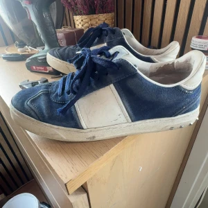 Valentino Garavani blå sneakers - Säljer ett par snygga Valentino Garavani sneakers i mörkblå mocka med vita detaljer och snörning. Skorna har en platt sula med coola mönster och små nitar på sidan. Perfekt för dig som gillar exklusiva märken och vill sticka ut med stil.