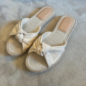 Beige sandaler med knut från H&M - Storlek 40, men små så passar 39!! Endast använda vid ett kort tillfälle! Snygga beige sandaler från H&M med öppen tå och platt sula. Ovandelen är i bomullstyg med en cool knutdetalj framtill. Sulan har en flätad kant som ger en chill vibe. Perfekta för varma dagar och enkel att slänga på sig till jeans eller klänning.