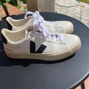 Vita sneakers från Veja med svart V - Snygga vita sneakers från Veja med svart V-logga på sidan och svart detalj vid hälen. Skorna har klassisk snörning och är tillverkade i skinn med rund tå och platt sula. Fint skick. 