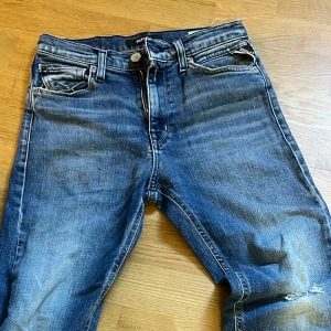 Replay blå jeans med slitningar - Säljer ett par snygga blå jeans från Replay med klassisk femficksdesign och slitna detaljer vid knä och benslut. Jeansen har rak passform och normal midja, med coola tvättade effekter och logopatch bak. Perfekt för dig som gillar en avslappnad streetstil.