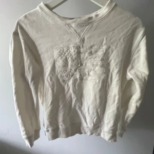 Vit sweatshirt från Polo Ralph Lauren - Säljer en vit sweatshirt från Polo Ralph Lauren i storlek M . Tröjan har ett broderat amerikanskt flaggmotiv på bröstet i samma färg som tyget. Rund hals och långa ärmar, tillverkad i mjuk bomull. Perfekt för dig som gillar clean och klassisk stil.140/152