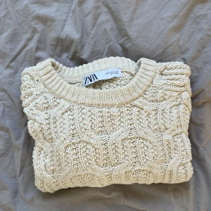 Beige stickad tröja från ZARA - Säljer en beige stickad tröja från ZARA med grovt flätmönster och rund hals. Tröjan har långa ärmar och svart kant längst ner för en snygg kontrast. Perfekt för dig som gillar chill och stilren vibe. Materialet är mjuk bomull som känns skönt mot huden.