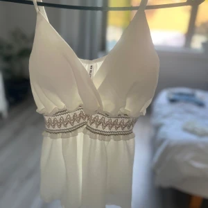 Vit omlott linnetopp från New Collection - Supersöt vit linnetopp med omlott-skärning och smala axelband i storlek S. Midjan har ett broderat zigzag-band i beige och ljusrosa toner. Toppen har inlägg och materialet känns som tunn polyester. Plagget är som ny, använd en gång. ordinarie pris är 200kr men säljer för 150kr