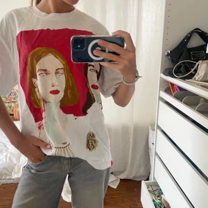 Vit t-shirt med konsttryck från Zara - Vit oversize t-shirt från Zara med stort konstnärligt tryck på framsidan som föreställer två ansikten mot en röd bakgrund. T-shirten har rund hals och korta ärmar. Materialet är mjuk bomull och passformen är loose. Perfekt för dig som gillar unika prints och vill sticka ut.