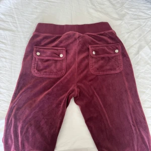Juicy Couture byxor  - Säljer ett par mjukisbyxor från Juicy Couture med bred resår i midjan och snörning framtill. Byxorna har två stora fickor med knappar bak och broderad JC-logga på ena fickan fram. Materialet är mjukt och skönt velour, perfekt för chill dagar.