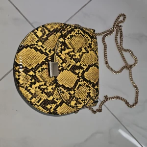 Gul axelväska med ormmönster - Snygg gul axelväska med svart ormmönster och guldfärgad kedja. Väskan har rundad form och ett metallspänne framtill. Perfekt för att lyfta din outfit med lite extra edge och färg.