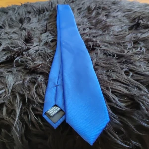 Klarblått polyester slips  - Snyggt och stilrent slips i klarblå färg, tillverkat av polyester. Bältet har en klassisk form och passar perfekt för att ge din outfit en färgklick. Enkelt att matcha med jeans eller chinos för en cool och ungdomlig look samt fungerar den även till  finare tillfällen 