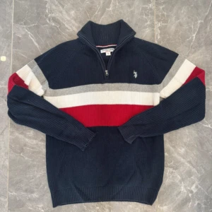 Randig stickad tröja från U.S. Polo Assn. - Snygg stickad tröja från U.S. Polo Assn. i marinblått, rött, grått och vitt. Tröjan har half zip, hög krage och långärmade ribbade muddar. Klassisk logga broderad på bröstet och coola breda ränder över hela tröjan. Perfekt för dig som gillar sportig stil.