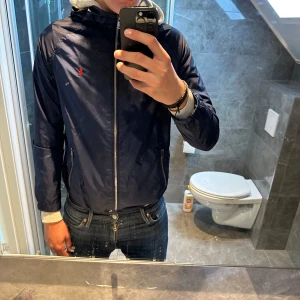Mörkblå vindjacka från Polo Ralph Lauren - Riktigt schysst mörkblå Ralph lauren regnjacka. Jackan är i storlek XL 18-20 vilket motsvarar en Xs/S. Jackan är i riktigt bra skicka utan några defekter. Modellen är 187cm