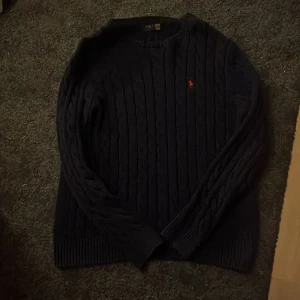 Mörkblå kabelstickad tröja Polo Ralph Lauren - Säljer en mörkblå kabelstickad tröja från Polo Ralph Lauren med röd broderad logga på bröstet. Tröjan har rund hals, långa ärmar och ribbade muddar. Perfekt för dig som gillar klassisk stil med en modern touch. Den sitter inte så tajt utan mer avslappnat. Säljer den pga att den e för stor