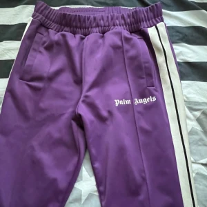 Palm Angels lila trackpants strl L - Säljer ett par snygga lila trackpants från Palm Angels i storlek L. Byxorna har vita och svarta sidoremsor, elastisk midja och Palm Angels-logga tryckt på benet. Materialet är polyester och de har en loose passform med raka ben. Perfekta för en chill streetwear-look.