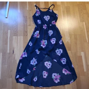Marinblå långklänning med blommor - Säljer en marinblå långklänning med blommigt mönster i rosa och lila toner. Klänningen har smala axelband och omlott-design framtill, vilket ger en snygg och flowy look. Perfekt för varma dagar och somriga dagar. 