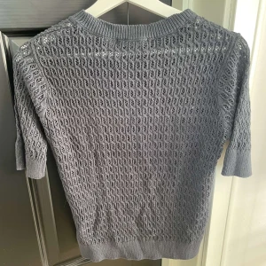 Grå stickad tröja från 157 - Snygg grå stickad tröja från 157 med korta ärmar och coolt hålmönster över hela plagget. Rund halsringning och ribbade muddar vid hals, ärmslut och nederkant. Perfekt för dig som gillar en chill och trendig look.