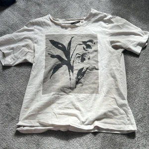 Vit t-shirt med blommigt tryck Lindex XS - Vit t-shirt från Lindex i storlek XS med ett stort svart blommigt tryck på framsidan. Klassisk rund hals och korta ärmar. Mjuk och skön bomull som passar perfekt till jeans eller shorts. Enkel och stilren design med en liten twist.