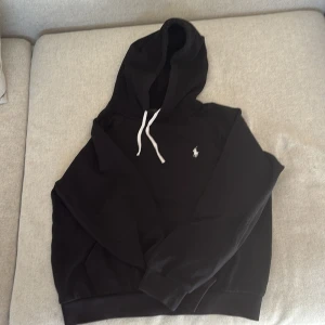Svart hoodie från Polo Ralph Lauren - Svart hoodie från Polo Ralph Lauren med vit broderad logga på bröstet och vita snören vid huvan. Tröjan har lång ärm, ribbade muddar och är gjord i mjuk bomull. Perfekt för chill dagar och enkel att matcha med jeans eller joggers. Säljer då den är för liten för mig. 