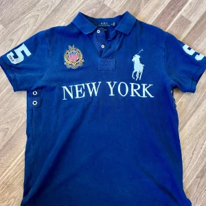 Blå Polo Ralph Lauren t-shirt - Snygg mörkblå t-shirt från Polo Ralph Lauren med klassisk polokrage och knappar. Stor vit logga med ryttare, broderad emblem och texten 'NEW YORK' framtill. Nummer 5 på båda ärmarna och coola detaljer vid sidorna. Perfekt för dig som gillar sportig stil.