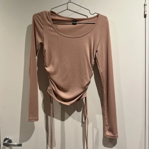 Ribbad beige långärmad topp - Säljer en beige ribbad topp i storlek M. Toppen har långa ärmar, rund halsringning och snygga snörningsdetaljer på sidorna som ger en croppad look.