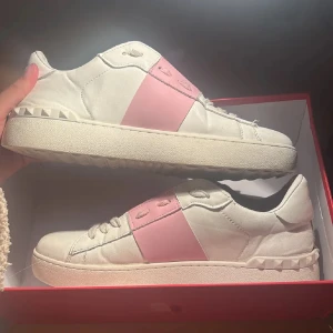 Valentino sneakers med rosa detalj - Snygga sneakers från Valentino Garavani i vitt skinn med bred rosa panel över snörningen. Skorna har platt sula med ikoniska nitar längs kanten och rund tå. Perfekt för dig som gillar lyxiga och trendiga detaljer. Äkta