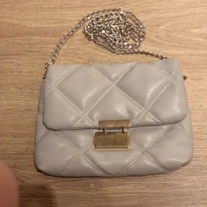 Beige quiltad axelväska med kedja - Snygg beige axelväska med quiltad design och guldfärgat lås framtill. Väskan har en silverfärgad kedja som axelrem och ett mjukt, lyxigt material. Perfekt för att lyfta din outfit med en stilren touch.