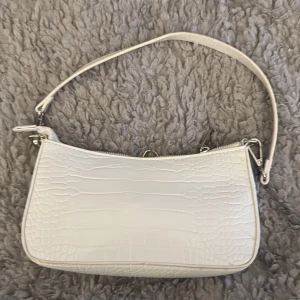 Vit axelväska med krokodilmönster - Snygg vit axelväska med krokodilmönstrad yta och silverfärgade detaljer. Väskan har en stilren, rektangulär form och ett justerbart axelband. Stängs med dragkedja upptill och passar perfekt för att lyfta din outfit med en trendig touch.