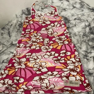 Blommig kortklänning från L.O.G.G. - Säljer en färgstark kortklänning från L.O.G.G. med blommigt mönster i rosa, vit, orange och brunt. Klänningen har halterneck och är ärmlös, perfekt för varma dagar. Materialet är mjuk bomull och den har en cool retro-vibe med tydlig logga nertill. Passar 140-160 (xxs-s)