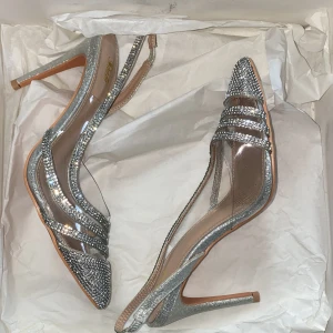 Glittriga pumps med spetsig tå - Säljer ett par glamorösa pumps från Shein med spetsig tå och hög klack. Skorna har transparenta paneler och är täckta med glittriga strassdetaljer i silver och guld. Slingback-modell med smal rem runt hälen och platt sula framtill. Perfekta för att sticka ut.