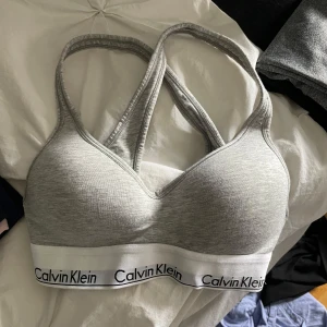 Sport bh från Calvin Klein - Supersnygg grå sportbh från Calvin Klein med sportig racerback och bred vit resår med logga. Mjuk och stretchig bomullsmix som sitter skönt mot kroppen. Perfekt för chill eller träning, och den har en clean look med diskret v-ringning framtill. 75b. Aldrig använd 