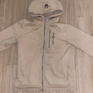 Beige hoodiejacka från Sail Racing - Beige hoodiejacka från Sail Racing med dragkedja framtill och på bröstfickan. Jackan har broderad logga på bröstet och huvan, samt två sidofickor. Mjuk och skön polyesterkvalitet, perfekt för chill dagar. Snygg och enkel design med lång ärm. Pris kan diskuteras 